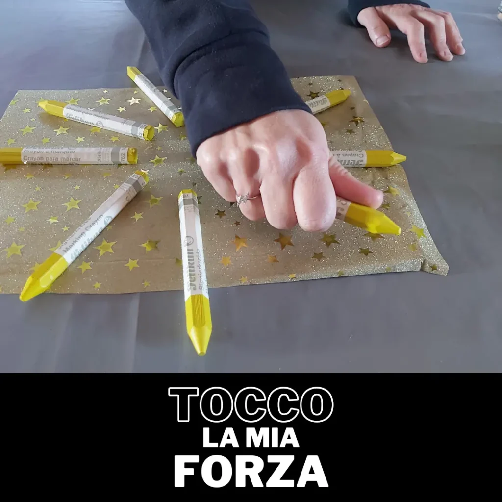 Primo piano di una mano che stringe un pennarello giallo, appoggiato con altri, su una carta regalo dorata. Scritta in basso 'TOCCO LA MIA FORZA'. Immagine usata per rappresentare la crescita personale in un laboratorio di arteterapia.