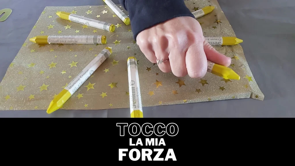 Primo piano di una mano che stringe un pennarello giallo, appoggiato con altri, su una carta regalo dorata. Scritta in basso 'TOCCO LA MIA FORZA'. Immagine usata per rappresentare la crescita personale in un laboratorio di arteterapia.